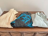Three sweaters folded on cabinet: beige cardigan, gray with teal pattern, mint green cardigan