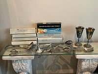 Full view of assorted silver-plated flatware, goblets and boxes on glass shelf