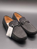 Three-quarter view of both loafers showing texture, metal bit, attached retail tag