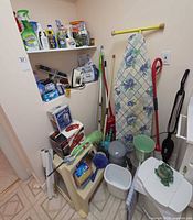 Overall view of closet area showing ironing board, Dirt Devil vac, organizer, cleaning products, trash bin and plant stand