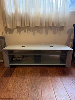 Front view of silver/light gray TV stand showing open shelf, glass shelf, cable holes, overall condition