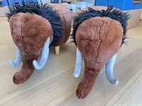 Front view of both plush mammoth ottomans showing tusks, manes and one wooden leg
