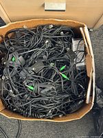 Box filled with assorted black cables and power adapters showing RJ11 cords, 3.5 mm green-tipped audio cables, barrel-plug DC leads, and wall bricks