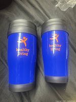 Two identical blue Healthy Living travel mugs standing upright showing logo and lids