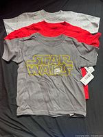 Front view of three stacked T-shirts showing gray logo tee on top with tags