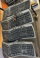 Four Microsoft ergonomic keyboards stacked, showing USB cables, key layout, and brand markings