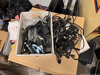 Tray containing assorted power adapters/bricks and mixed cables alongside large pile of black power cords