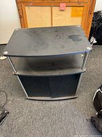 Front angled view of the black engineered wood TV stand showing three tiers, glass doors, and metal posts