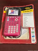 Front view of TI-84 Plus CE calculator in blister pack showing pink unit, keypad, color screen graphic, included USB cable