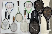 Overview of all nine rackets laid on table showing variety of colors, head shapes and one cover
