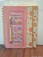 Notebook front with accessory pack overlaid, showing floral cover, spiral binding, pen and sticker sheet