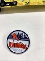 Front view of round enamel Raiders hockey pin with tape measure for scale