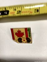 Front of square enamel pin showing maple leaf and soccer ball design with tape measure for scale