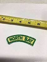 Front view of curved green and yellow North Bay patch beside tape measure showing size