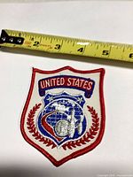 Full view of embroidered United States shield patch with tape measure for scale