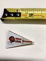 Front of triangular enamel pin beside ruler showing approx. 1.5 in. length