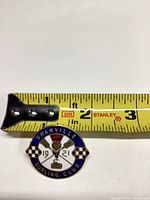 front view of enamel pin beside tape for scale, shows design and 1921 date