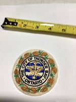 Front view of round Thunder Bay Ontario embroidered patch beside tape for scale