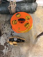 Orange reel with yellow extension cord beside rolled green tarp tied with rope and multi-tool in holster