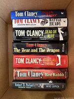 Stack of seven hardcover thriller novels with visible spine titles