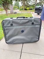 Front view of gray Santosha suitcase showing external zip pocket, logo tag, webbing straps and handle