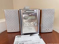 Front view of JVC main unit flanked by two speakers, manual visible
