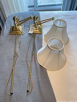 Pair of brass wall-mount swing arm lamps with wiring and two off-white fabric shades displayed on bed