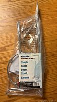 Front view of sealed package showing label 'Simple Houseware Simple Toilet Paper Stand, Chrome' with metal parts visible