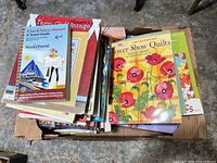 Box containing stack of various craft books and magazines, top book Flower Show Quilts visible