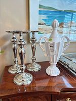 Three silver metal candlesticks and white lidded urn on tabletop