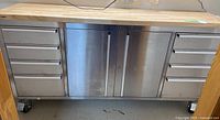 Front view of stainless steel workbench showing drawers, cabinet doors, butcher block top and casters