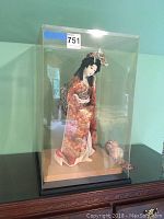 Front view of the Japanese Geisha doll in a clear display case on a wooden surface.