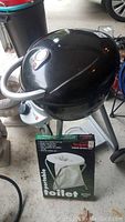 Front and side view of the black CharBroil propane grill on its stand, with a portable toilet box in front.