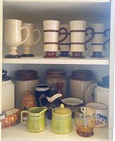 cabinet shelves showing crocks, canisters, mugs, pitchers, soup bowl, glass