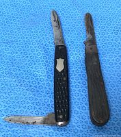 Two folding pocket knives open showing blades, black synthetic handle knife with shield and wood handle knife