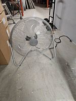 Front view of 20-inch metal floor fan on concrete floor; shows blades, grille, base and power cord.
