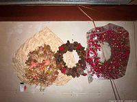 Overview showing three wreaths hung on wall: dried flower on straw base, pinecone with berries, plastic berry wreath in bag