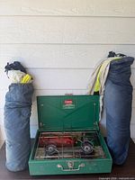 Stove with lid open showing burners and fuel tank flanked by two folded chairs in bags