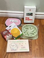 Group shot showing all five pet items: diaper box, booties box, dental wipe box, green slow feeder bowl, yellow treat toy
