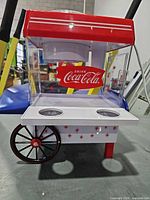 Front view of red and white Nostalgia Coca-Cola snow cone machine showing cup holders and wheel