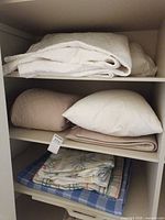 Closet shelves with folded white comforter, beige comforter, pillow, floral bedding and blue plaid blanket