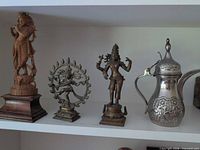 Four items on shelf: carved wood Krishna, Shiva Nataraja statue, Ardhanarishvara statue, silver-plated Dallah pot