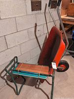 Folded Corrie kneeler bench beside red wheelbarrow against concrete wall