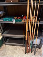 Shelf with five cues, boxed Aramith ball set, loose extra balls, triangle rack and accessories visible