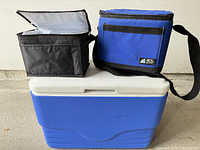 Coleman cooler closed with two lunch bags on top, black tote open