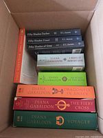 Box showing eleven visible paperback books including Diana Gabaldon titles, E. L. James series, and Derek Hansen spine