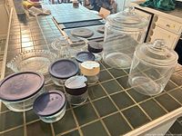 Overview of all glass items: two canisters, three blue-lidded Pyrex bowls, four spice jars, pie dish, cruet