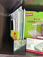 Left shelf side showing metal organizer with assorted notebooks and Staples 9 x 12 envelope box in background