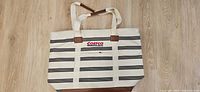 Front view of cream canvas Costco tote with black stripes, brown leather bottom, handles and shoulder strap visible
