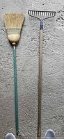 Straw broom and metal fan rake laid on concrete showing full length and condition